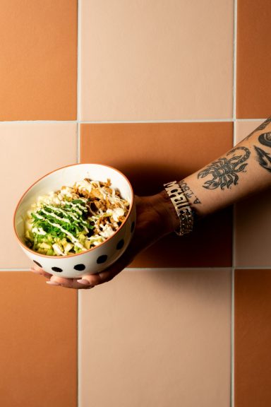 A hand holding a bowl of rice with green vegetables against a tile background.